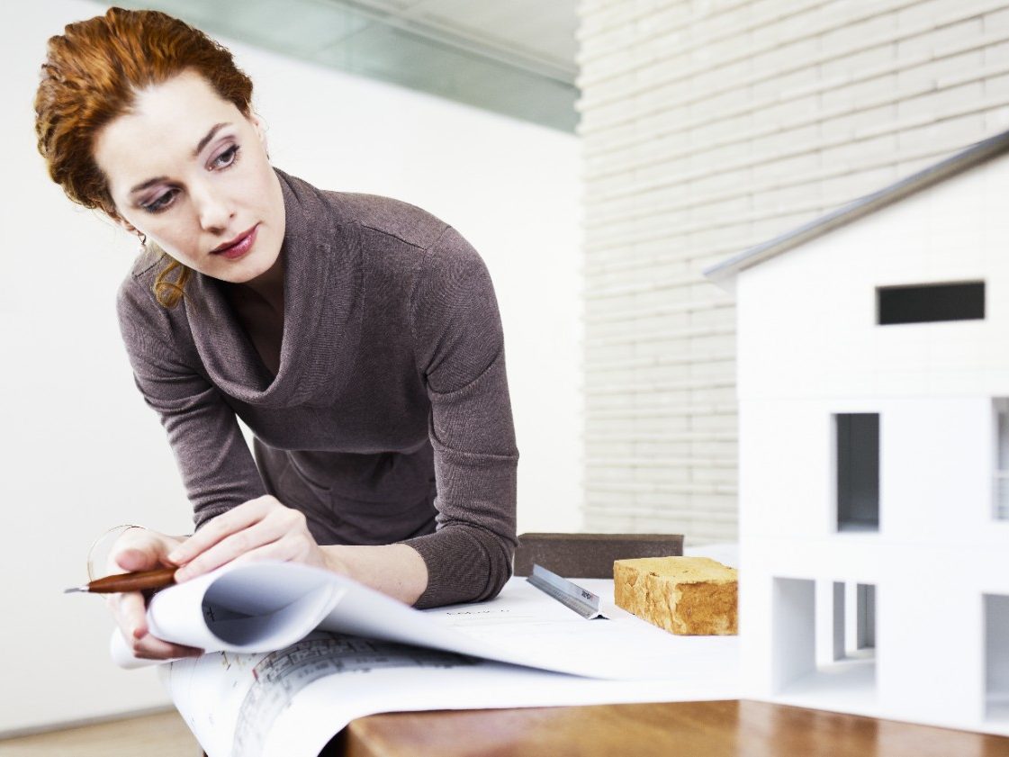 Female planner or architect with blueprint and mock-up of a house at the office