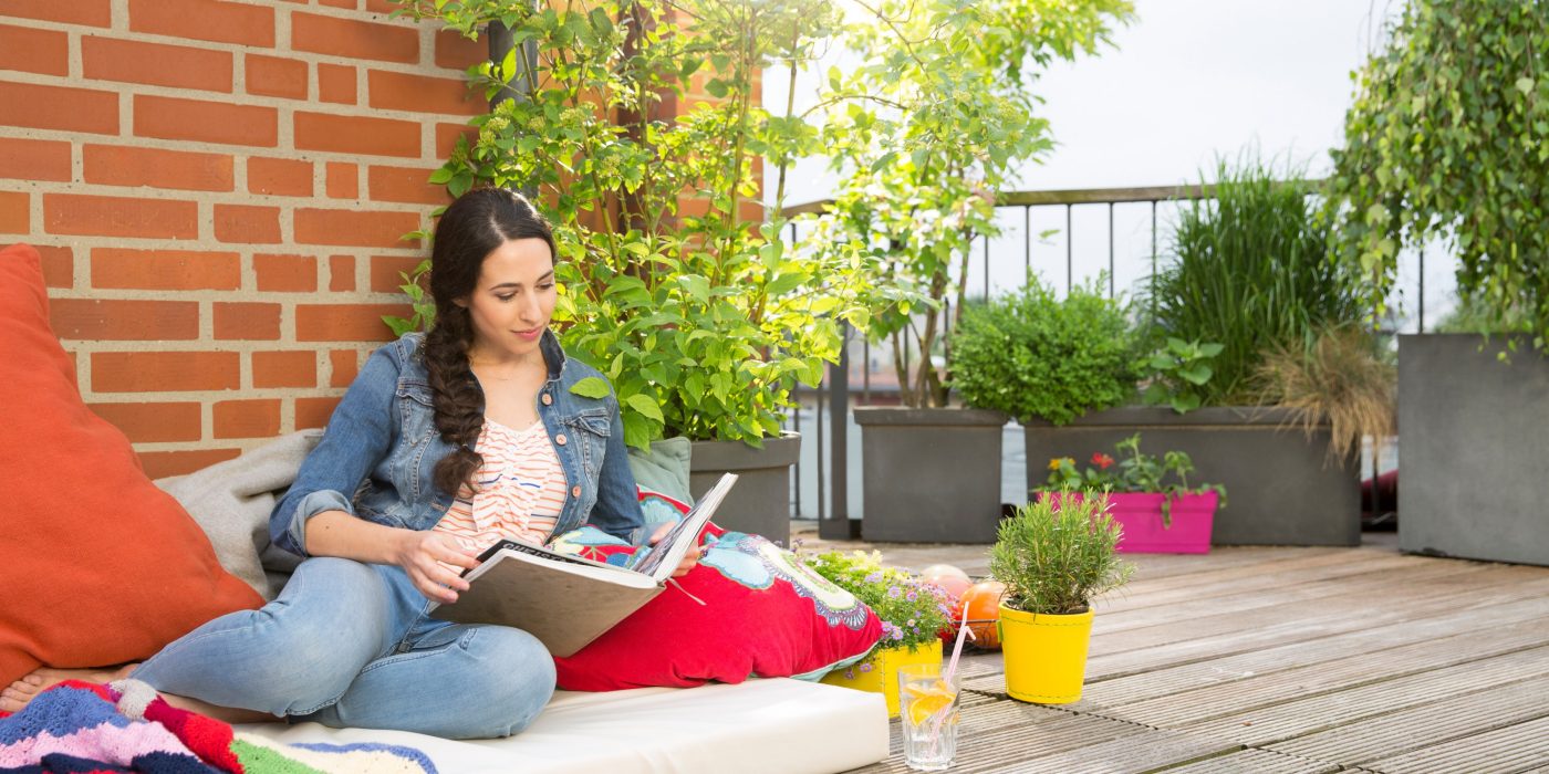 Woman surrounded by colorful cushions and blankets on rooftop terrace flipping through photo album in front of brick wall