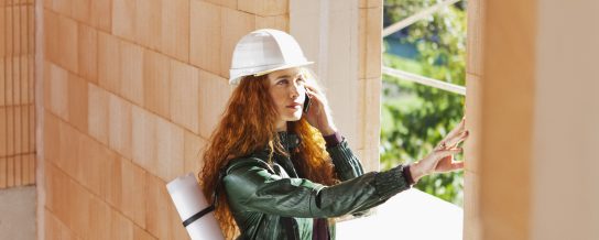 Female architect with hard hat and blueprint-casing inside a building shell touching a brick